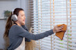 Excited young asian woman with headphones singing and dancing while dusting a window in living room, well-decorated room, joyful woman singing while cleaning in living room, lifestyle concept.