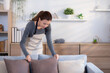 Young asian woman diligent housekeeper arrange cushions on sofa in living room at home, woman wearing apron doing housework and cleaning pillow on couch, lifestyle and domestic life concept.