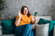 Hi. Brunette taking a selfie photo using her mobile phone at home. 30s woman in yellow t-shirt recording self video, talking on online call and relaxing on a sofa.