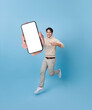 Full length of cheerfull Asian man jumping and smiling in air with showing mobile phone blank screen with empty space for mobile app on screen isolated on blue background.