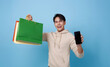 Trendy young Asian handsome man carrying colorful bags shopping online with mobile phone mockup of blank screen isolated on blue background.