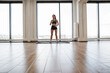 © sofiko14 - Fit young woman wearing workout gear stands on mat in spacious gym with large windows overlooking the city. Sports woman with dumbbells, ready for exercise routine, embodying health and fitness.