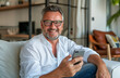 © Nordic - Close up smiling man wearing glasses using smartphone, sitting on couch at home, happy male looking at phone screen, browsing apps, chatting or shopping online, having fun with gadget