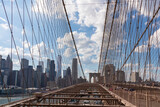 Fototapeta Góry - The gate of Brooklyn Bridge with a waving American flag on top of it contrasted with a blue sky with puffy, white clouds. Suspension bridge in New York City. Numerous skyscrapers in the back.