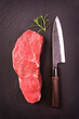 Raw dry aged angus roast beef steak offered as top view  on a design board with a Japanese santoku knife