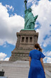 A woman in blue dress standing under an iconic representation of freedom and independence, the Statue of Liberty with flaming torch on Liberty Island. The Lady on a Pedestal is surrounded by clouds.