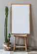 A blank white poster in an oak frame leaning against the wall on top of a small wooden stool, a cactus plant beside it, neutral colors, natural lighting, and soft shadows.