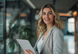 © PedroEnrique - Portrait of a smiling business woman standing in a modern office with a laptop and tablet, looking at the camera. The female manager is holding a digital device and working on a project or social