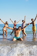 Vertical Portrait of a group of happy young people in swimming costumes posing for a photo on the shore of the beach. Diverse friends smiling and making playful gestures raising arms looking at camera