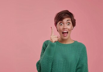 Wall Mural - Photo of an excited woman in a green sweater pointing her finger up at empty space isolated on a pastel pink background. A happy young female with short hair showing a blank area for text while