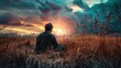 A person is sitting in a field of tall, golden grass, looking towards a dramatic sunset with rich orange and blue tones in the sky. Clouds are scattered across the sky, and patches of sunlight are bre
