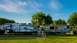 Fifth wheel trailers parked back to back at campsite on grass with trees and blue cloudy skies