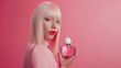 A blonde girl with make-up is holding a pink bottle in her hands on a pink background. Advertises perfume. Place for text.