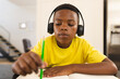 An African American boy wearing headphones, writing in notebook at home