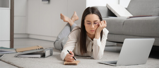 Tired young businesswoman lying on floor with laptop and notebook at home