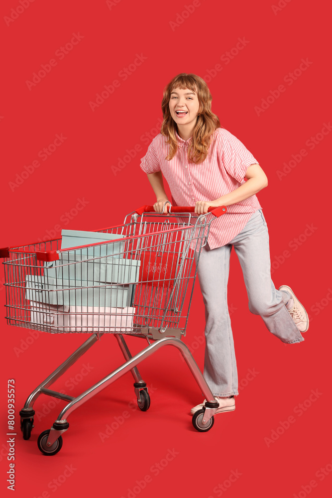 Young woman with boxes in shopping cart on red background