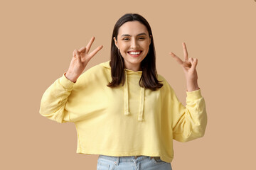 Sticker - Smiling young woman showing victory gesture on brown background