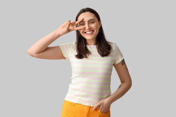 Sticker - Smiling young woman showing victory gesture on grey background