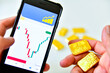 Hand holding a smartphone and a gold bar to check the gold price graph for investment