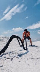 Wall Mural - Topless man working out with a rope on nature. Muscular athlete exercising with battle ropes on the white mountain against blue sky in summer. High intensive workout. Vertical video