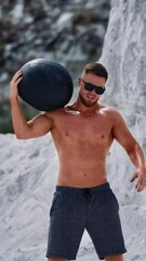 Wall Mural - Bodybuilder training with a heavy ball in white mountains. Strong guy wearing black sunglasses doing exercises with a ball over his head outdoors in summer. Vertical video