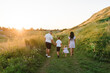Mother father holding hands baby daughter, son walk in mountains at sunset. Happy family spending time together on summer day outdoors. Children with parents walking in green grass in field. Back view