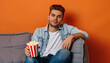 Handsome young man with popcorn watching TV on grey sofa against orange background