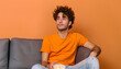Handsome young man with popcorn watching TV on grey sofa against orange background