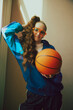 Young girl in orange glasses, colorful, vintage-inspired blue tracksuit, striking makeup and curly hair posing with basketball ball. Concept of 90s, fashion, youth culture, old-style trends