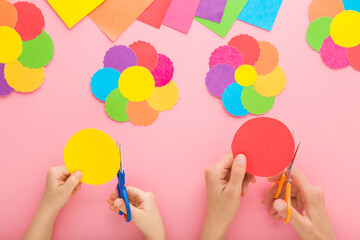 Wall Mural - Young adult mother and child hands holding scissors, cutting paper and making colorful flower shapes on light pink table background. Pastel color. Closeup. Creating decoration elements. Top down view.