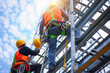 © samsusam - Two workers wearing safety helmets and orange work vests, with green posture tape tied around their waists, are climbing up from ground level view at a steel structure roof on a sunny day.