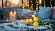 Soft pillows, blanket, burning candles and yellow tulips on rattan garden furniture in evening