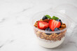 Tasty granola with berries and yogurt in plastic cup on white marble table, closeup. Space for text
