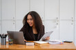 © Wasana - Stressed African American businesswoman reviewing documents while working on a laptop in a modern office.
