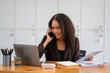 © Wasana - Cheerful African American businesswoman having a phone conversation while working on a laptop in a modern office.
