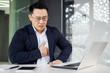 © Liubomir - An Asian businessman appears distressed, clutching his chest in pain while sitting at a desk in a modern office. He looks at his laptop screen with a worried expression.