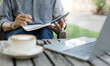 A man taking a note in book use laptop working outdoor with coffee drink on old wooden table
