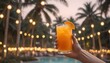 A hand holding a glass of orange cocktail in front of a blurred tropical background with palm trees and string lights