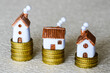 Three different small toy houses on yellow coin stacks of different heights, different cost concept