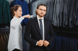Handsome man in suit, beautiful female consultant helps removes dust and hair from clothes in classic menswear store