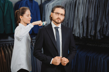 Wall Mural - Handsome man in suit, beautiful female consultant helps removes dust and hair from clothes in classic menswear store