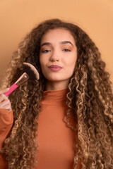 confident young woman using make up brush in beige studio background. beauty, care, fashion concept.