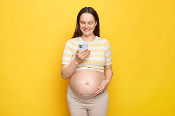 Wall Mural - Overjoyed Caucasian pregnant woman with bare belly wearing casual top isolated over yellow background holding smartphone checking social networks chatting online