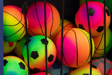 A Bin Of Colorful Toy Soccer And Basketballs In A Wire Bin For Sale. The Balls Are Pink, Purple, Yellow, Green And Yellow Colored. The Soccer Balls Have Black Pentagon Shapes And The Balls Have Lines.