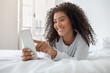 A cheerful young Hispanic woman with curly hair is lying on a white bedspread, engrossed in using her smartphone. The natural light from the rooms window casts a warm glow