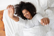 Hispanic woman is lying on a bed covered with white sheets, stretching arms. She appears comfortable and relaxed, with her eyes closed. The room is simple and clean, with minimal decoration.