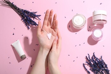 Wall Mural - Woman applying hand cream and lavender flowers on pink background, top view