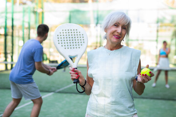 Wall Mural - Portrait of positive elderly woman standing on padel tennis court, holding racket and ball and smiling.