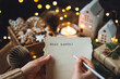 Christmas letter on craft paper to Santa Claus with text: Dear Santa. Cozy home interior with candles, lights, handmade toys, gingerbread cookies. Wishlist for presents from a child or adult