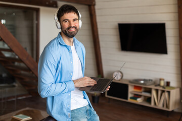 Poster - A man wearing a blue shirt is standing and holding a laptop in his hands. He appears focused on the screen, possibly working or browsing from home, using headphones